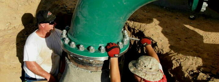Two workers adjusting and installing a green, curved pipe within a construction site.