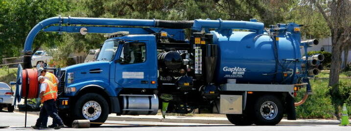 A vacuum truck, with a large hose, is seen in use on a street, possibly for cleaning or maintaining pipes.